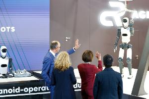 Bundeskanzler Friedrich Merz winkt einem Roboter zu – die Zukunft winkt zurück.
