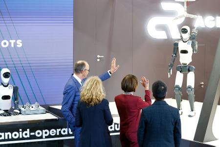 Bundeskanzler Friedrich Merz winkt einem Roboter zu – die Zukunft winkt zurück.