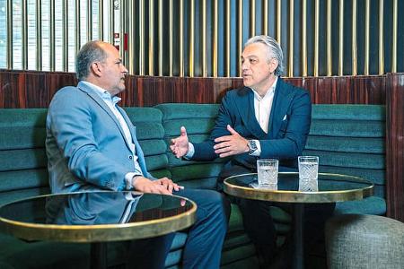 Luca de Meo und Redakteur Stefan Cerchez sitzen in einer Lounge auf einem grünen Sofa und führen ein Gespräch. Auf den Tischen vor ihnen stehen Gläser mit Wasser. Die Szene wirkt wie ein Interview oder Geschäftsgespräch in gehobenem Ambiente.