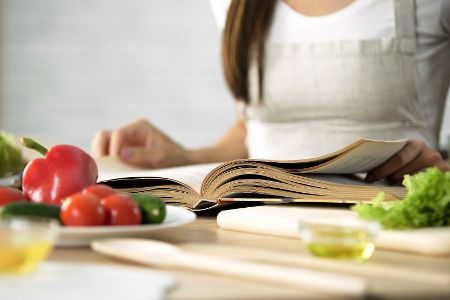 Junge Frau steht zwischen Kochzutaten vor einem Rezeptbuch