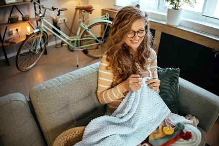 Junge Frau sitzt in ihrer Wohnung und häkelt eine Decke