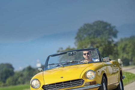 Gelber Fiat 124 Spider Cabrio auf Landstraße, frontal fotografiert, Fahrer am Steuer, sommerliche Landschaft.
