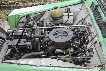 Detailaufnahme eines Motorraums eines älteren Fahrzeugs mit sichtbarem Vergasermotor, Batterie und Kühlwasserbehälter.