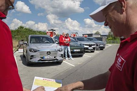 Vergleichstest von vier Elektro-SUVs (BMW i5 30xDrive, Mercedes GLC 400 4MATIC EQ, Audi Q6 SUV e-tron quattro, Porsche Macan 4S) an einer Ladestation mit mehreren Personen im Vordergrund.