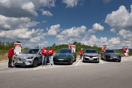 Vier Elektro-SUVs – BMW i5 30xDrive, Mercedes GLC 400 4MATIC EQ, Audi Q6 SUV e-tron quattro und Porsche Macan 4S – stehen nebeneinander an Ladesäulen auf einem Parkplatz, begleitet von mehreren Personen in roten Oberteilen, die Messungen oder Notizen durchführen. Der Himmel ist leicht bewölkt.