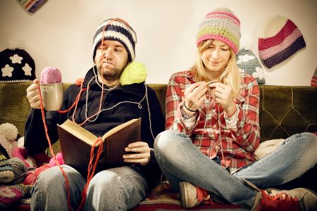 Junge Paar sitzt auf dem Sofa, wo sie inmitten von Wollchaos strickt und er ein Buch liest