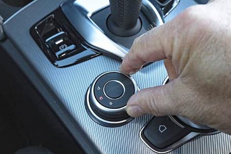 Detailaufnahme des Drehreglers und der Mittelkonsole im Alfa Stelvio 2.0 Diesel. Die Hand eines Fahrers bedient den Fahrmodusschalter neben dem Automatik-Wählhebel.