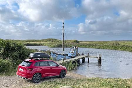 Skoda Karoq 2.0 TDI mit Dachbox an einem kleinen Bootsanleger in Dänemark, im Vordergrund ein rotes SUV, im Hintergrund ein Boot am Steg und weite Wiesenlandschaft unter bewölktem Himmel.