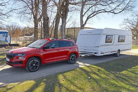 Ein roter Skoda Karoq zieht einen weißen Dethleffs Camper Wohnwagen auf einem Campingplatz. Im Hintergrund sind Bäume und ein bunt bemalter Bauwagen zu sehen. Die Aufnahme entstand bei Tageslicht im Frühjahr.