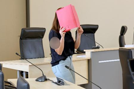 Die Angeklagte betritt den Gerichtssaal mit rotem Aktendeckel vor dem Gesicht.