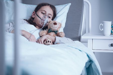 Kind mit Atemmaske und Teddy im Arm liegt im Krankenhausbett