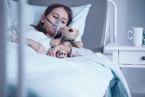 Kind mit Atemmaske und Teddy im Arm liegt im Krankenhausbett