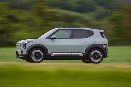 Der Dateiname 'ams_Kia EV2_RG26_061.jpg' gibt eindeutig das Modell 'Kia EV2' an. Das Bild zeigt einen hellgrauen Kia EV2 SUV in Seitenansicht, fahrend auf einer Landstraße mit unscharfem, grünem Hintergrund. Es handelt sich um eine Fotografie eines Elektro-SUVs im Fahrbetrieb.