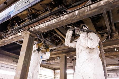 Mitarbeiter der Werkstatt der Verkehrsaktiengesellschaft VAG zeigen, wie ein Trockeneisstrahlgerät angesetzt werden muss, um ein durch Bitum verschmutztes Fahrwerks einer Straßenbahn zu reinigen (gestellte Szene). 