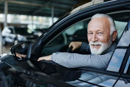 Ein älterer Mann mit weißem Bart am Steuer eines Pkw schaut lächelnd aus einem geöffneten Autofenster
