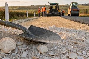 Eine Straßenbaustelle in der Froschperspektive. Vorne eine Schaufel die auf einem Haufen weißen Muschelpulver liegt, teilweise sind noch Jacobsmuscheln zu erkennen. Im Hintergrund asphaltieren Bauarbeiter eine Straße.
