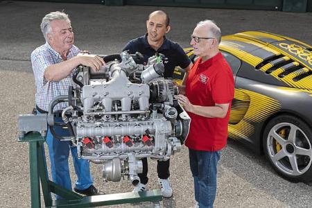 Drei Personen stehen um einen Ruf 8Zylinder Prototyp-Motor, der auf einem Motorständer montiert ist. Im Hintergrund ist ein Sportwagen mit auffälliger gelb-schwarzer Folierung zu sehen.