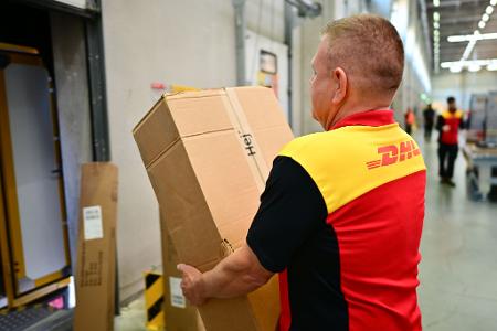 Ein Paketbote schleppt in einem DHL-Standort ein schweres Paket. (Archivbild)