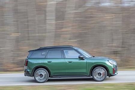 Mini JCW Countryman 4all fährt auf einer Landstraße, Seitenansicht, grüne Lackierung, unscharfer Waldhintergrund.