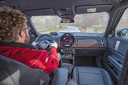 Innenraumaufnahme eines Mini JCW Countryman 4all während der Fahrt, Blick über die Schulter des Fahrers auf das Cockpit und die Straße.