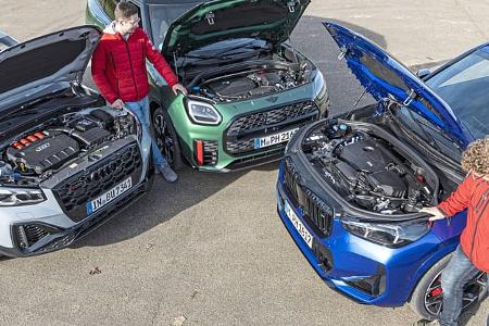 Drei Kompakt-SUVs – Audi SQ2, BMW X1 M35i xDrive und Mini JCW Countryman – mit geöffneten Motorhauben, zwei Personen betrachten die Motoren.