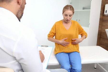 Frau sitzt beim Arzt, hält sich den Bauch und erklärt ihre Symptome