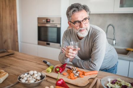 Älterer Mann steht in der Küche beim Gemüse schneiden und trinkt ein Glas Wasser