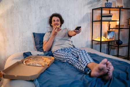 Mann liegt im Bett mit der Fernbedienung in der Hand und isst Pizza