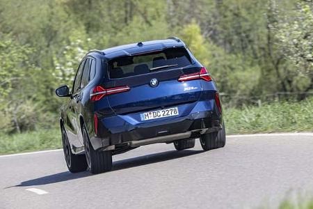 BMW X3 20 xDrive fährt auf einer Landstraße durch eine grüne, bewaldete Landschaft. Das Fahrzeug ist von hinten zu sehen.