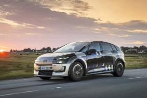 Audi A2 e-tron Erlkönig (Tarnfahrzeug) fährt auf Landstraße bei Sonnenuntergang. Das Fahrzeug trägt eine auffällige Tarnfolierung, im Hintergrund ist eine ländliche Szenerie mit Häusern und Wiesen zu sehen.