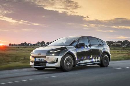 Audi A2 e-tron Erlkönig (Tarnfahrzeug) fährt auf Landstraße bei Sonnenuntergang. Das Fahrzeug trägt eine auffällige Tarnfolierung, im Hintergrund ist eine ländliche Szenerie mit Häusern und Wiesen zu sehen.