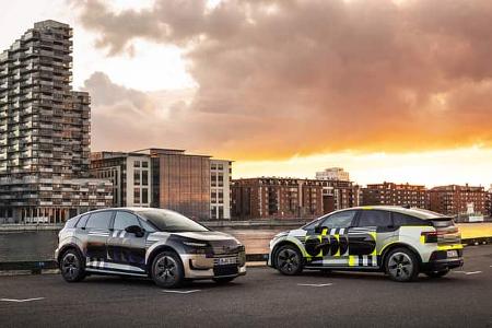 Zwei Audi A2 e-tron Erlkönig Prototypen auf einem Parkplatz vor moderner Stadtarchitektur bei Sonnenuntergang.
