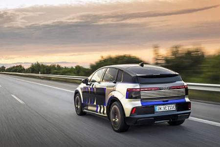 Audi A2 e-tron Erlkönig auf offener Straße bei Sonnenuntergang, getarntes Elektroauto in Fahrt.