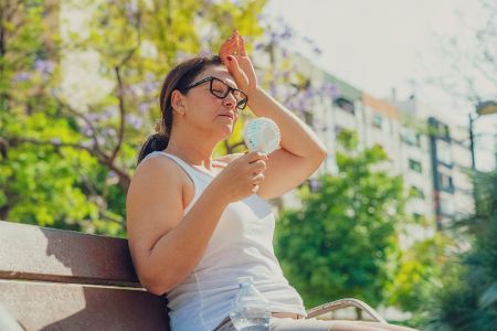 Frau sitzt mit Ventilator auf Parkbank und schwitzt