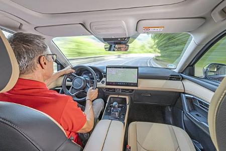 Innenaufnahme eines Mazda CX-5 2,5l E-Skyactive G während der Fahrt, Blick über die Schulter des Fahrers auf das Cockpit und die Straße.