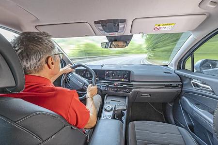 Innenaufnahme eines Kia Sportage 1,6 T-GDI während der Fahrt, Blick über die Schulter des Fahrers auf das Cockpit und die Straße.