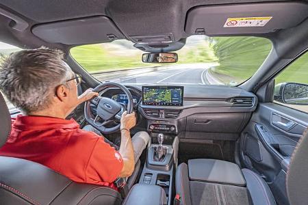 Innenraumaufnahme eines Ford Kuga 1,5l EcoBoost während der Fahrt, Blick aus der Rücksitzperspektive auf Fahrer, Cockpit und Armaturenbrett.