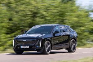 Cadillac Lyriq-V fährt auf einer Landstraße, Seitenansicht, schwarze Lackierung, grüne Vegetation im Hintergrund.