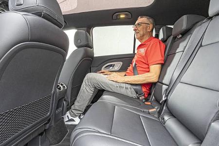 Innenaufnahme eines Mercedes GLB 250+ mit Fokus auf die Rücksitzbank. Ein Mann sitzt auf der Rückbank und demonstriert die Beinfreiheit und Sitzposition. Die Lederausstattung und das Panoramadach sind sichtbar.