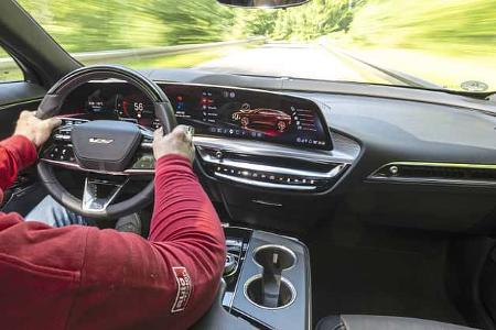 Innenaufnahme eines Cadillac Lyriq-V während der Fahrt, Blick über die Schulter des Fahrers auf das digitale Cockpit und die Straße.