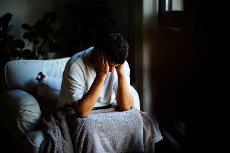 Depressiver Mann sitzt mit Decke auf der Couch und hat sein Gesicht in den Händen vergraben