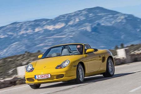 Gelber Porsche Boxster 986 Cabrio fährt auf einer kurvigen Bergstraße, im Hintergrund Gebirge, Fahrer am Steuer.