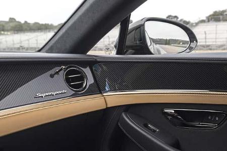 Die Aufnahme zeigt das Interieur der Beifahrertür eines Bentley Supersports mit hochglänzender Carbonfaser-Zierleiste und dem „Supersports“-Schriftzug. Sichtbar sind runde Luftdüse, Chrom-Türgriff, Verriegelungstasten sowie zweifarbige Lederverkleidung mit Kontrastnähten. Durch das Fenster und den Außenspiegel ist die Umgebung einer Rennstrecke im Hintergrund erkennbar.