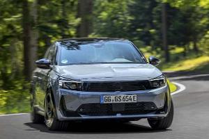 Das Foto zeigt einen Opel der Kompaktklasse in Frontansicht, der eine Kurve auf einer Landstraße durch bewaldetes Gebiet fährt. Die markante Opel‑Vizor‑Front mit schmalen Scheinwerfern und sportlicher Schürze ist erkennbar; das deutsche E‑Kennzeichen deutet auf einen Elektro- oder Plug‑in‑Hybridantrieb hin. Der Wagen ist dynamisch in Bewegung abgebildet.