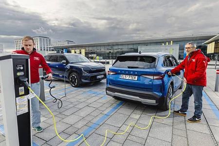 Zwei Elektroautos, ein Kia EV3 und ein Skoda Elroq, werden an einer Ladesäule auf einem Parkplatz geladen. Zwei Personen in roten Jacken bedienen die Ladevorgänge. Im Hintergrund ist ein modernes Gebäude zu sehen, der Himmel ist bewölkt.