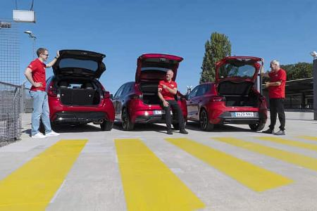 Drei rote Kleinwagen – Renault Clio, Seat Ibiza und Toyota Yaris Hybrid – stehen nebeneinander mit geöffneten Heckklappen auf einem Parkplatz. Drei Personen in roten Shirts interagieren jeweils mit den Fahrzeugen. Die Szene zeigt eine Testumgebung bei sonnigem Wetter.
