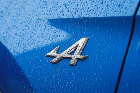 Die Detailaufnahme zeigt das Markenemblem der Alpine A390 GT auf einer blauen Karosserieoberfläche. Das verchromte Alpine‑A‑Logo ist von zahlreichen Wassertropfen bedeckt und hebt die Konturen sowie die Spaltmaße der angrenzenden Karosserieteile hervor.