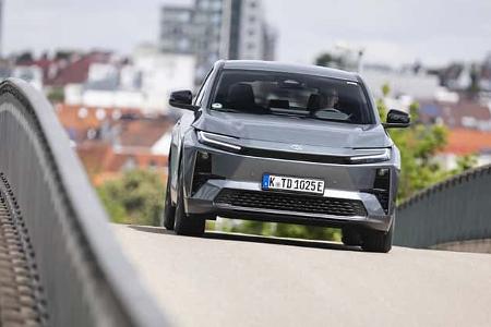 Toyota C-HR+ BEV auf einer Brücke in urbaner Umgebung, frontal fotografiert, moderne Architektur im Hintergrund.