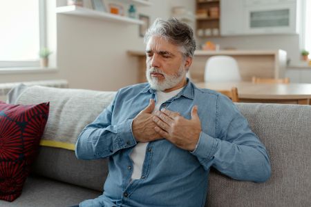 Älterer Mann sitzt auf dem Sofa und hält sich die schmerzende Brust