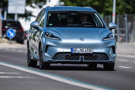 Das Foto zeigt den MG4 Urban in der Frontansicht während der Fahrt im Stadtverkehr. Der vollelektrische Kompaktwagen besitzt eine weitgehend geschlossene Front mit markant integrierten LED-Scheinwerfern und einer aerodynamisch gestalteten Stoßfängerpartie. Die Aufnahme erfolgt auf einer öffentlichen Straße in Deutschland, erkennbar am E-Kennzeichen.